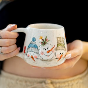 Könnte beinhalten: Keramikbecher mit drei lächelnden Schneemännern mit Karottennasen und Wintermützen. Die Schneemänner sind auf einem gesprenkelten weißen Hintergrund gemalt. Der Becher hat einen großen Henkel und wird von einer Person gehalten.