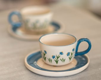 May include: Two ceramic teacups and saucers. The cups are white with blue handles and rims, and decorated with blue flower and green stem designs. The saucers are white with a blue border. The background is a light gray surface.