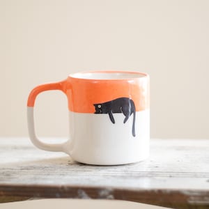 Könnte beinhalten: Eine Keramik-Tasse mit einem skurrilen Design. Die obere Hälfte ist orange, die untere Hälfte ist weiß, und eine schwarze Katzenillustration befindet sich auf dem weißen Bereich. Der Griff ist orange und grau. Eine lustige und einzigartige Kaffeetasse.