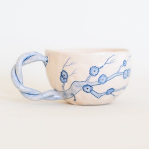 May include: A handmade ceramic teacup with a twisted blue handle. The off-white cup features a delicate blue floral branch design. The cup is a unique piece of pottery, perfect for enjoying tea or coffee.