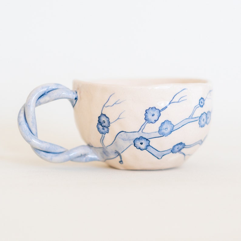 May include: A handmade ceramic teacup with a twisted blue handle. The off-white cup features a delicate blue floral branch design. The cup is a unique piece of pottery, perfect for enjoying tea or coffee.