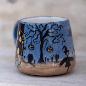 May include: A ceramic Halloween-themed mug with a blue background and a spooky scene. The design includes a haunted house, a bare tree with jack-o'-lanterns, a scarecrow, and tombstones. The mug has a light blue handle and a tan base.