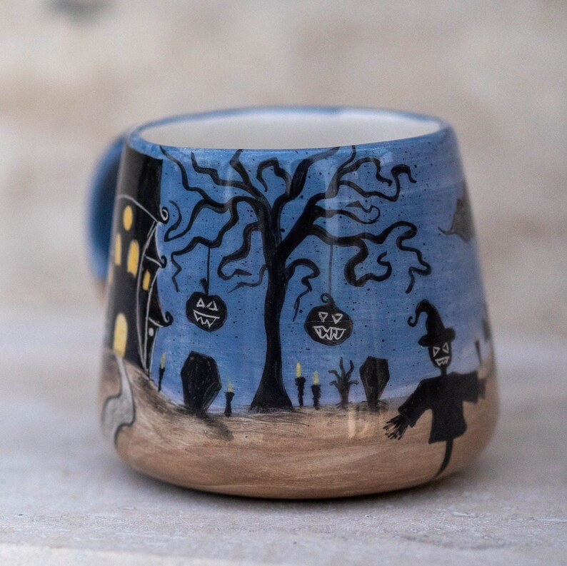 May include: A ceramic Halloween-themed mug with a blue background and a spooky scene. The design includes a haunted house, a bare tree with jack-o'-lanterns, a scarecrow, and tombstones. The mug has a light blue handle and a tan base.