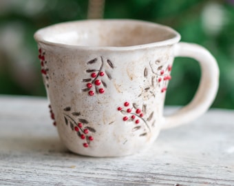 May include: Cream-colored ceramic mug with a handle, decorated with dark brown leaf and red berry designs. The mug sits on a white wooden surface, with a blurred green background. The mug has a rustic, handmade appearance.