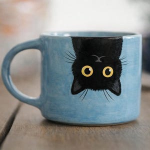 Op de afbeelding: Een lichtblauwe keramische mok met een zwart kattenontwerp. De kat is ondersteboven afgebeeld met gele ogen en snorharen. De mok heeft een gebogen oor en staat op een houten oppervlak.