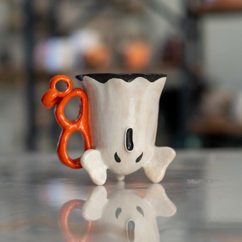K&ouml;nnte beinhalten: Eine wei&szlig;e, keramikartige, geisterf&ouml;rmige Tasse mit einem orangefarbenen Griff. Die Tasse hat einen schwarzen Rand und schwarze Details f&uuml;r Augen und Mund. Die Tasse steht auf einer reflektierenden Oberfl&auml;che.