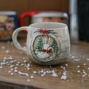 May include: A ceramic mug with a winter scene design. The mug has a handle and a round image of a snowy landscape with a house, trees, and a snowman. A red bow decorates the top of the image. The mug is on a wooden surface.