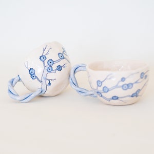 May include: Two ceramic mugs with a cream-colored base and blue floral designs. The handles are twisted and painted blue. One mug is upright, the other is tilted on its side. The mugs are on a white surface.