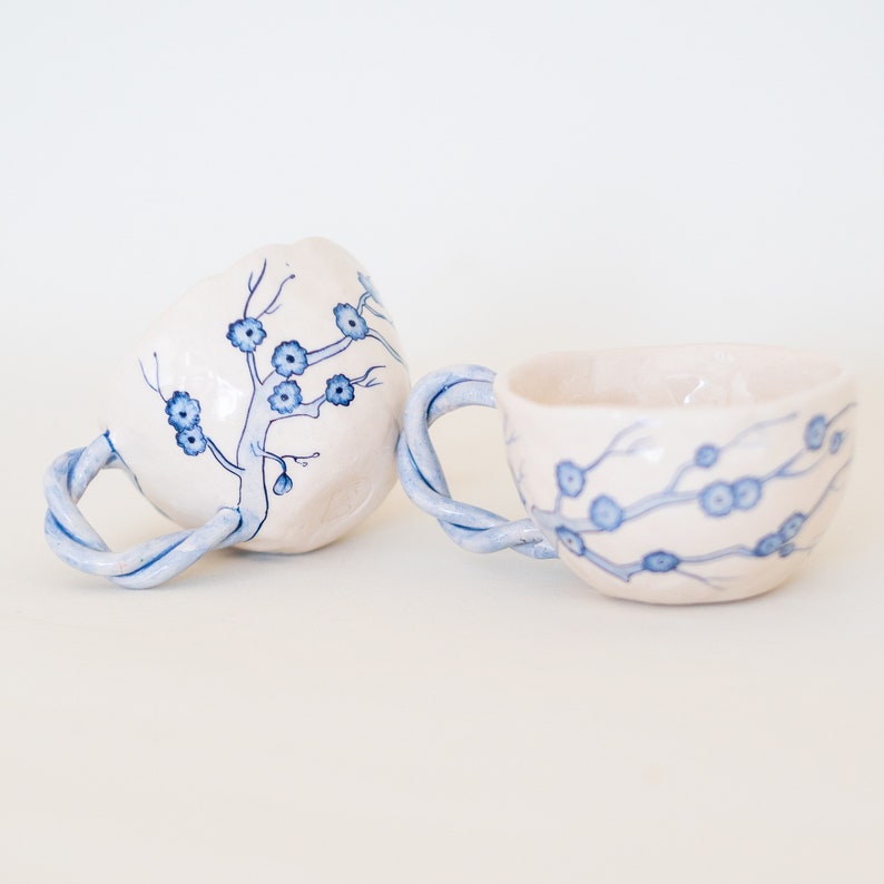 May include: Two ceramic mugs with a cream-colored base and blue floral designs. The handles are twisted and painted blue. One mug is upright, the other is tilted on its side. The mugs are on a white surface.