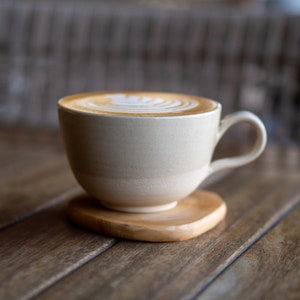 May include: A beige ceramic coffee cup with a handle, filled with a latte featuring a decorative foam design. The cup sits on a small, natural wood coaster. The background is a wooden surface.