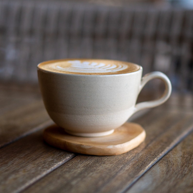 May include: A beige ceramic coffee cup with a handle, filled with a latte featuring a decorative foam design. The cup sits on a small, natural wood coaster. The background is a wooden surface.