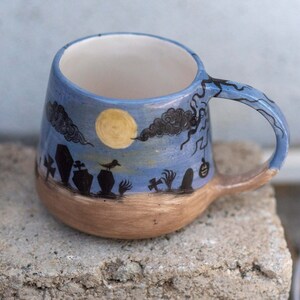 May include: A ceramic mug with a Halloween theme. The mug features a blue sky with a full moon, black clouds, and a graveyard scene. The handle and rim are blue, and the base is brown.