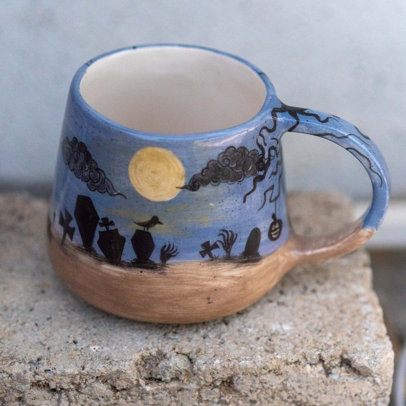May include: A ceramic mug with a Halloween theme. The mug features a blue sky with a full moon, black clouds, and a graveyard scene. The handle and rim are blue, and the base is brown.