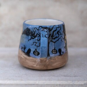 May include: A ceramic Halloween mug with a blue background and a spooky tree design. The mug features black tree branches, jack-o'-lanterns, and a graveyard scene. The bottom of the mug is a light brown color.