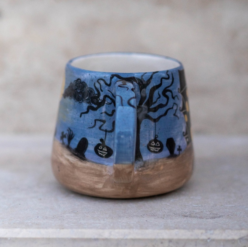 May include: A ceramic Halloween mug with a blue background and a spooky tree design. The mug features black tree branches, jack-o'-lanterns, and a graveyard scene. The bottom of the mug is a light brown color.