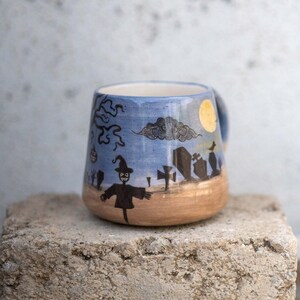 May include: A ceramic Halloween-themed mug with a blue sky background and a graveyard scene. The mug features a scarecrow, tombstones, a full moon, and a tree. The bottom of the mug is tan.