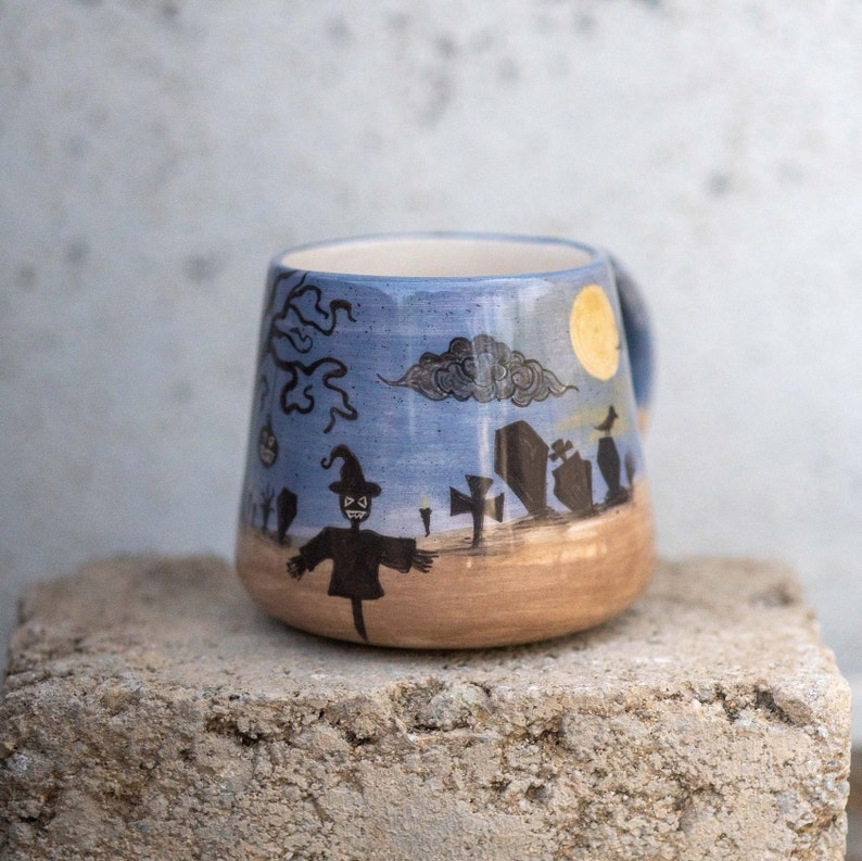 May include: A ceramic Halloween-themed mug with a blue sky background and a graveyard scene. The mug features a scarecrow, tombstones, a full moon, and a tree. The bottom of the mug is tan.