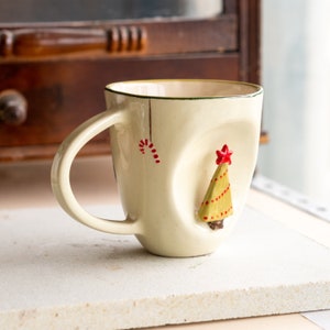 Puede incluir: Taza de cerámica color crema con diseño navideño. La taza presenta un árbol de Navidad amarillo en 3D con adornos de puntos rojos y una estrella roja. Un bastón de caramelo rojo y blanco está pintado en el lateral.