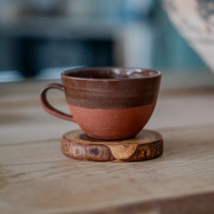 May include: A two-tone ceramic coffee cup, dark brown at the top and reddish-brown below, with a curved handle. It rests on a small, round wooden coaster. The cup and coaster are placed on a light-coloured wooden surface.