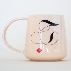 May include: A blush-colored ceramic mug with a curved handle. The mug features a black abstract design with a red diamond accent. The design is a modern, minimalist style, suitable for coffee or tea.