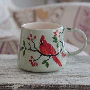 May include: A ceramic mug with a light green glaze, featuring a hand-painted red cardinal bird perched on a branch with red berries and green leaves. The mug has a rounded shape and a curved handle.