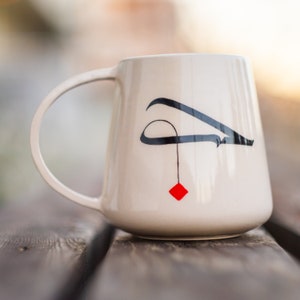 May include: Cream ceramic mug featuring a black abstract design and a small red square. The mug has a large handle and is placed on a wooden surface. A simple, modern design for tea or coffee.