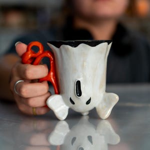 K&ouml;nnte beinhalten: Handgefertigte Keramiktasse in Geisterform. Die wei&szlig;e Tasse hat einen schwarzen Rand und Mund sowie einen orangefarbenen Griff. Die Tasse ist so konzipiert, dass sie auf zwei kleinen F&uuml;&szlig;en steht. Einzigartiges Design.