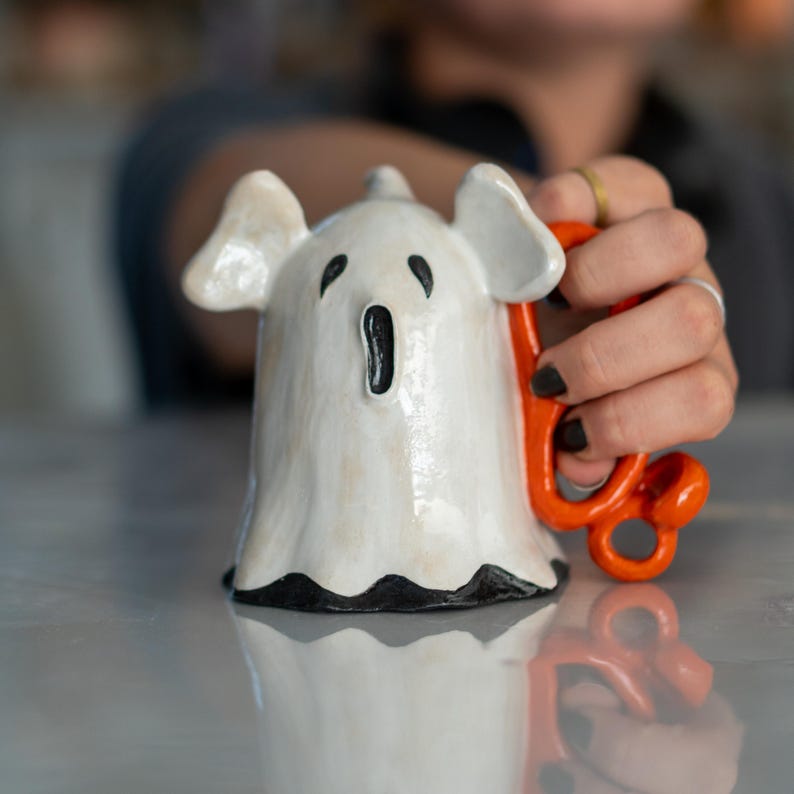 K&ouml;nnte beinhalten: Eine wei&szlig;e Keramik-Geister-Tasse mit einem orangefarbenen Griff. Der Geist hat schwarze Augen und einen weit ge&ouml;ffneten Mund. Der Boden der Tasse hat einen schwarzen Rand. Die Tasse wird von einer Person gehalten.
