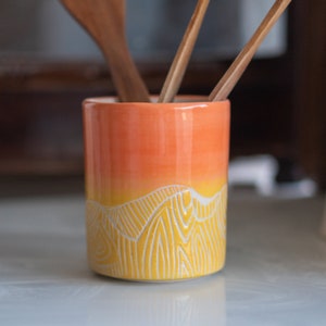 Puede incluir: Un portautensilios de cerámica cilíndrico con un degradado de naranja a amarillo. La parte inferior presenta un diseño de montaña blanco grabado. Se ven cucharas de madera en el interior. El soporte está sobre una superficie blanca.