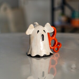 K&ouml;nnte beinhalten: Keramik-Geisterfigur mit wei&szlig;em K&ouml;rper, schwarzen Augen und Mund sowie einem orangefarbenen "BOO"-Anhang. Der Geist hat ein leichtes Used-Finish und einen schwarzen Besatz am unteren Rand, ideal f&uuml;r Halloween-Dekorationen.