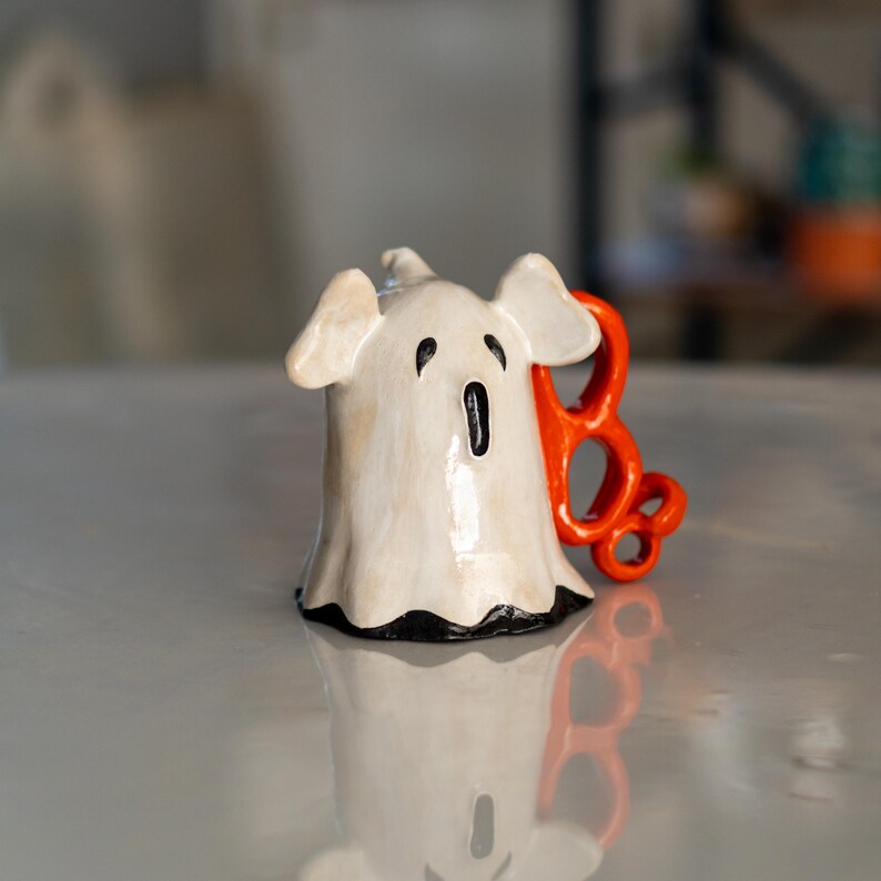 K&ouml;nnte beinhalten: Keramik-Geisterfigur mit wei&szlig;em K&ouml;rper, schwarzen Augen und Mund sowie einem orangefarbenen "BOO"-Anhang. Der Geist hat ein leichtes Used-Finish und einen schwarzen Besatz am unteren Rand, ideal f&uuml;r Halloween-Dekorationen.
