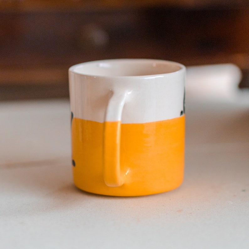May include: A small ceramic mug with a white upper half and a bright yellow lower half. The mug has a curved handle and a simple design. The interior is white. The mug is likely used for drinking coffee or tea.