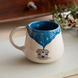 Op de afbeelding: Een crèmekleurige keramische mok met een blauwe binnenkant. De blauwe binnenkant heeft een sterrennachtontwerp dat naar beneden druipt naar een kleine huisillustratie op de voorkant. De mok heeft een afgeronde vorm en een gebogen handvat.