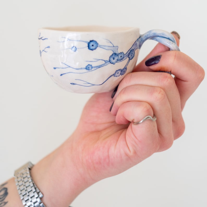 May include: A white ceramic teacup with a blue floral design. The handle and the floral branches are painted in blue. The cup is held in a hand against a white background. The cup is approximately 3 inches in diameter.