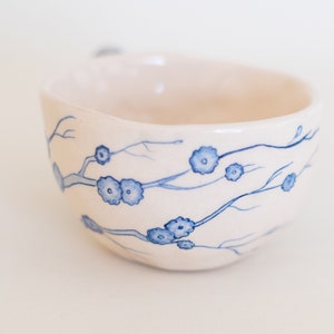 May include: A white ceramic bowl with a blue floral design. The bowl features a hand-painted pattern of delicate blue flowers and branches. The interior of the bowl is a soft white, and the exterior has a slightly textured surface.