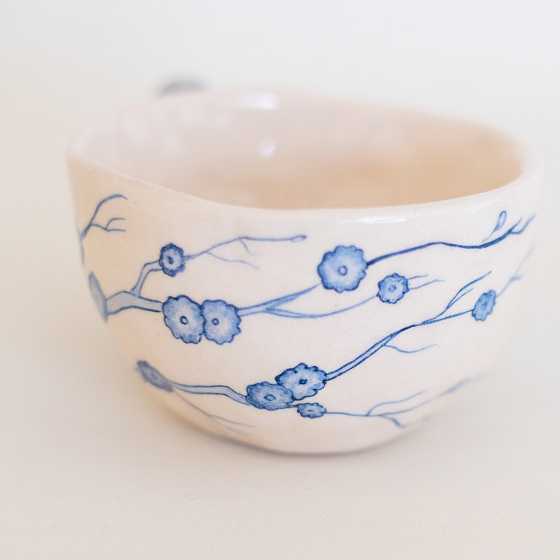 May include: A white ceramic bowl with a blue floral design. The bowl features a hand-painted pattern of delicate blue flowers and branches. The interior of the bowl is a soft white, and the exterior has a slightly textured surface.