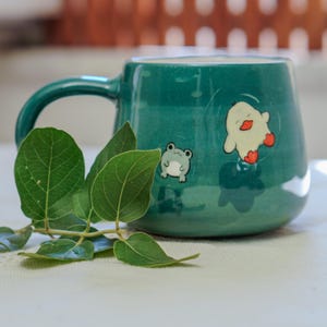 Könnte beinhalten: Eine türkisfarbene Keramiktasse mit abgerundeter Form und großem Henkel. Die Tasse zeigt Cartoon-Illustrationen eines Frosches und einer Ente. Grüne Blätter befinden sich im Vordergrund. Die Tasse steht auf einer weißen Oberfläche.
