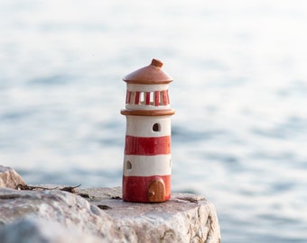 May include: A ceramic lighthouse with red and white stripes, a brown roof, and small windows. The decorative lighthouse sits on a rock with the ocean in the background. The lighthouse is a nautical-themed home decor item.