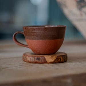 May include: A two-tone ceramic coffee cup, dark brown at the top and reddish-brown below, resting on a small, round wooden coaster. The cup features a curved handle. The cup is designed for hot drinks and is handmade.