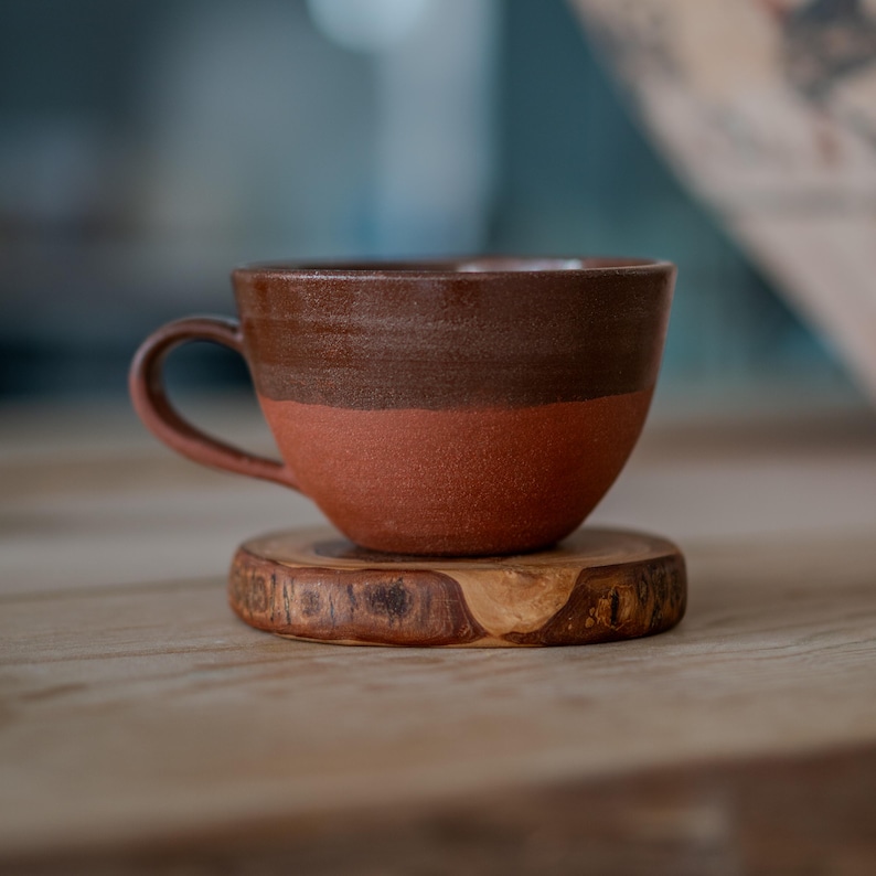 May include: A two-tone ceramic coffee cup, dark brown at the top and reddish-brown below, resting on a small, round wooden coaster. The cup features a curved handle. The cup is designed for hot drinks and is handmade.