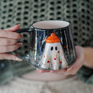 May include: A ceramic mug with a black exterior and a white ghost design. The ghost wears an orange hat and is decorated with red and yellow accents. The mug has a black handle and a white interior.