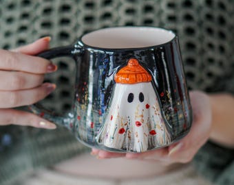 May include: A ceramic mug with a black exterior and a white ghost design. The ghost wears an orange hat and is decorated with red and yellow accents. The mug has a black handle and a white interior.