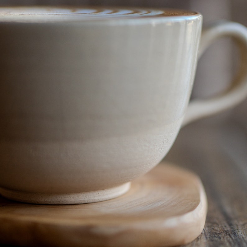 May include: A close-up of a beige ceramic coffee cup with a handle, filled with a latte, resting on a wooden coaster. The latte has a decorative pattern on top. The cup and coaster are on a wooden surface.