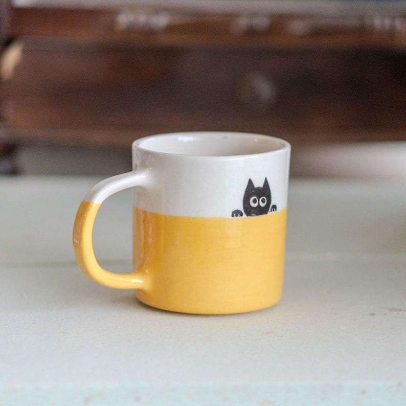 May include: A ceramic mug with a white upper half and a yellow lower half. A black cat illustration peeks over the yellow section. The handle is also yellow. The mug is on a white surface.