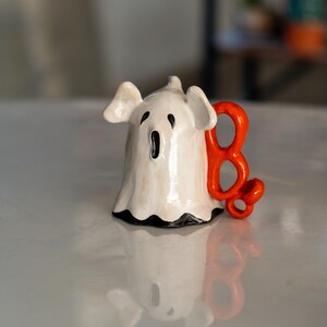 K&ouml;nnte beinhalten: Eine wei&szlig;e Keramik-Geisterfigur mit schwarzem Mund und Augen und einem leuchtend orangefarbenen "B" daran. Der Geist hat einen ger&uuml;schten unteren Rand und kleine Ohren. Eine Halloween-Dekoration.