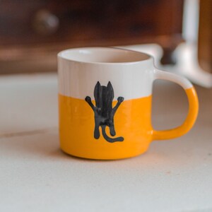 May include: A ceramic mug with a white top, a yellow middle, and a white handle with yellow accents. A black cat illustration is on the yellow section. The mug is designed for hot beverages.