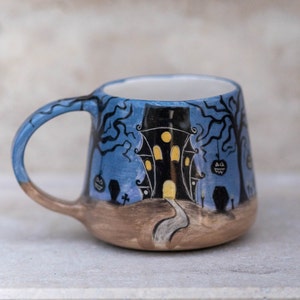 May include: A ceramic Halloween mug featuring a spooky haunted house design. The mug has a blue background with black trees, pumpkins, and a pathway leading to the house. The handle is blue and brown.