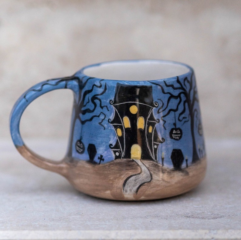 May include: A ceramic Halloween mug featuring a spooky haunted house design. The mug has a blue background with black trees, pumpkins, and a pathway leading to the house. The handle is blue and brown.