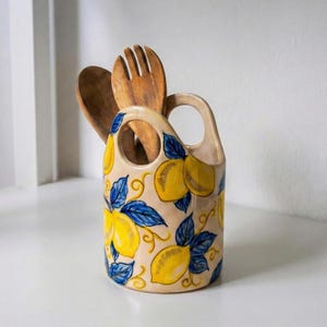 May include: A ceramic utensil holder with a handle, decorated with yellow lemons and blue leaves. Two wooden cooking utensils are inside. The holder is beige with a glossy finish, perfect for kitchen decor.