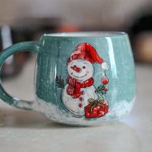 Könnte beinhalten: Eine türkisfarbene Keramiktasse mit einem skurrilen Schneemann-Design. Der Schneemann trägt einen roten Hut und Schal und hält eine rote Kugel. Eine rote Geschenkbox ist ebenfalls abgebildet. Die Tasse hat einen weißen, schneebedeckten Boden und einen passenden türkisfarbenen Henkel.